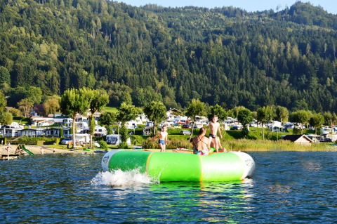 Bambini giocano su un grande gonfiabile in acqua vicino a un villaggio vacanze con glamping e roulotte.