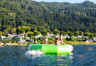 Kids playing on a large inflatable in the water near a holiday park with glamping and caravans in the background.