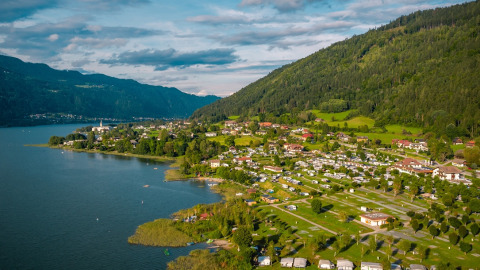 Vista aérea de un parque vacacional con glamping junto al lago, rodeado de colinas verdes y casas pequeñas.