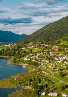 Vista aérea de un parque vacacional con glamping junto al lago, rodeado de colinas verdes y casas pequeñas.