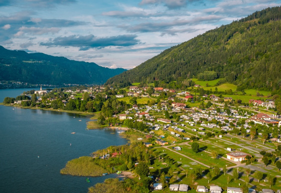 Luftaufnahme eines Ferienparks mit Glamping am See, umgeben von grünen Hügeln und kleinen Häusern.