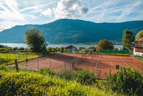 Cancha de tenis junto a un lago y montañas, día soleado, entorno natural en un parque vacacional.