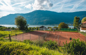 Tennisbane ved sø og bjerge i baggrunden, solrig dag i naturskønne omgivelser ved en feriepark.