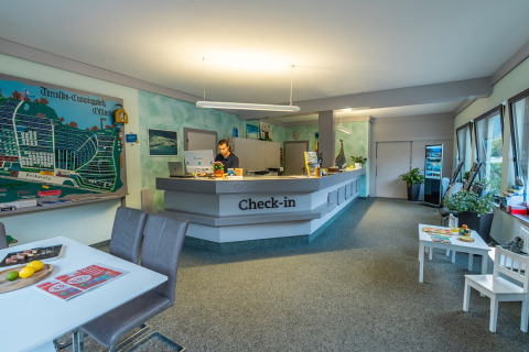 Mostrador de check-in en parque vacacional con glamping, empleado atendiendo, gran mapa del lugar.
