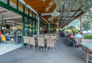Udendørs caféområde ved feriepark med glasfacade, grønne planter og gæster, ideelt til glamping.