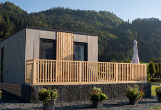 Moderne glampinghytte med træterrasse og bjerglandskab i baggrunden, beliggende i en feriepark.