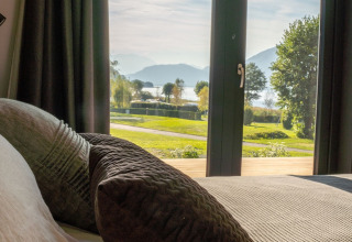 View from a glamping accommodation with large windows and cozy pillows, overlooking a lake and mountains.