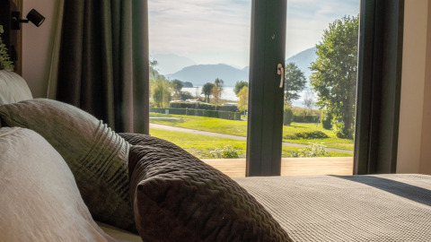 Vista desde un alojamiento glamping con grandes ventanas y cómodos cojines hacia lago, césped y montañas.