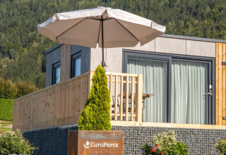 Moderne glampingaccommodatie met terras en parasol in een vakantiepark, omgeven door groene heuvels.