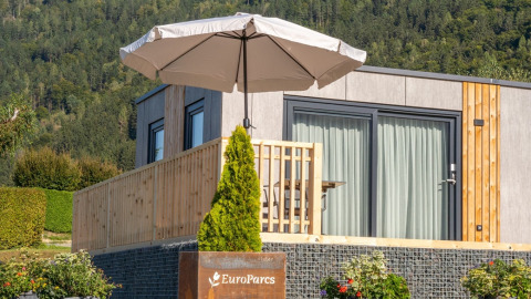 Moderne glamping-lodge met terras en parasol in een vakantiepark, omringd door bosrijke heuvels.