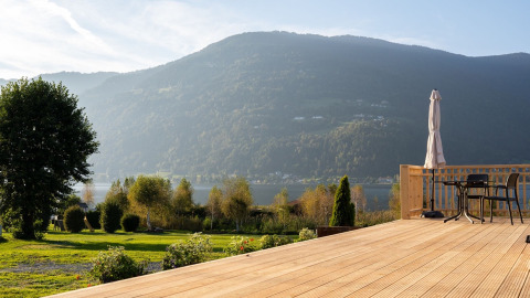 Zicht vanaf een houten terras met stoelen over een groen veld, bergen en meer bij een glampingpark.