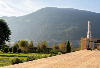 View from a wooden deck with chairs overlooking a green field, mountains and a lake at a glamping park.