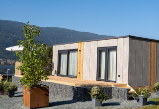 Moderne glamping lodge met houten terras en bloemrijke bakken in een vakantiepark met zicht op bergen.