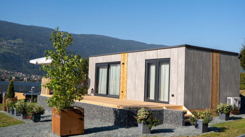 Modern glamping cabin with wooden deck and flower pots at a holiday park with scenic mountain backdrop.