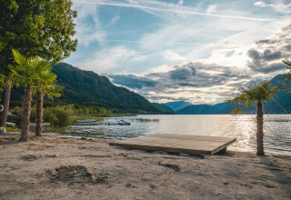 Beautiful lakeside view with mountains, sandy beach, and palm trees at a holiday park offering glamping options.