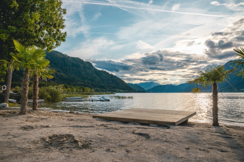 Hermosa vista al lago con montañas, playa de arena y palmeras en un parque vacacional con glamping.