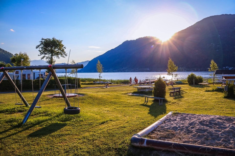 Parque de vacaciones soleado junto al lago con zona de juegos y montañas, perfecto para alojamiento glamping.