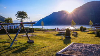 Parque de vacaciones soleado junto al lago con zona de juegos y montañas, perfecto para alojamiento glamping.