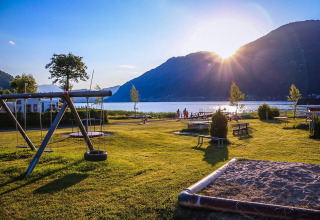 Solrig feriepark ved søen med legeplads, sandkasse og bjerge i baggrunden, ideel til glampingoplevelser.