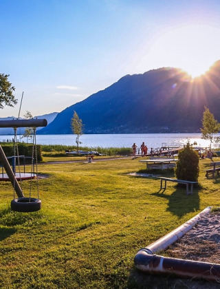 Parque de vacaciones soleado junto al lago con zona de juegos y montañas, perfecto para alojamiento glamping.
