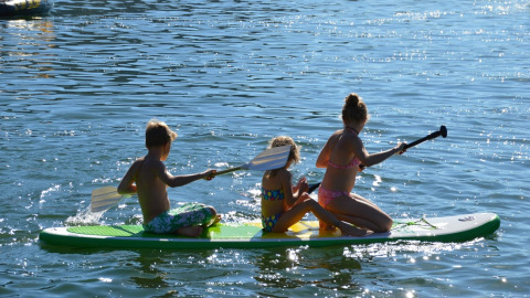 Tres niños reman juntos en una tabla de paddle surf en un lago en un parque vacacional con glamping.