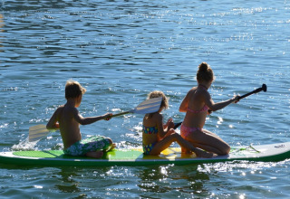 Tre børn padler sammen på et SUP-board på en sø ved en feriepark med glamping indkvartering.