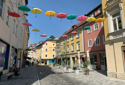 Sombrillas de colores cuelgan sobre una calle peatonal con edificios coloridos en un día soleado.