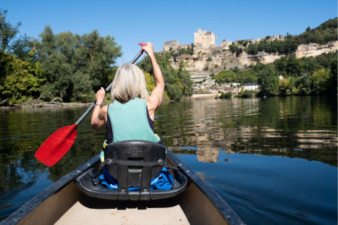 Mujer remando en canoa por un río tranquilo con árboles y un castillo al fondo en un parque de vacaciones.