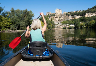 Woman paddling a canoe on a calm river, with a tree-lined shore and castle in the background at a holiday park.
