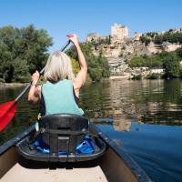 Mujer remando en canoa por un río tranquilo con árboles y un castillo al fondo en un parque de vacaciones.