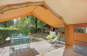 View from inside a safari tent, showing a deck with chairs, table, and trees in the background.