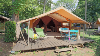 Tienda safari con terraza de madera, sillas verdes y mesa azul en un camping rodeado de árboles.