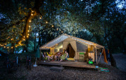 Four people relax outside a lit safari tent in the woods at night, surrounded by festive string lights.