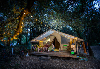 Four people relax outside a lit safari tent in the woods at night, surrounded by festive string lights.