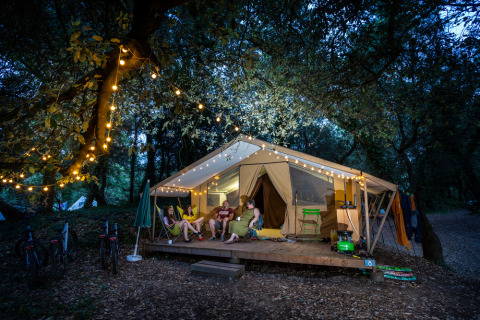 Cuatro personas descansan frente a una tienda safari iluminada en el bosque por la noche con luces.