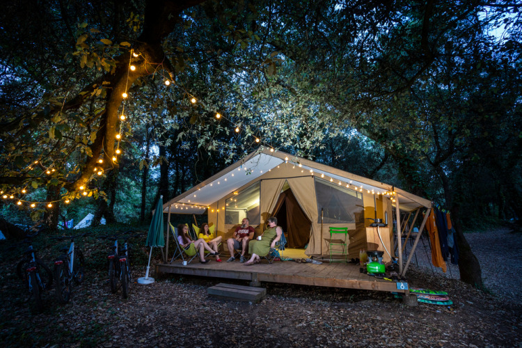 Four people relax outside a lit safari tent in the woods at night, surrounded by festive string lights.