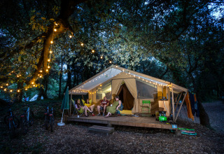 Four people relax outside a lit safari tent in the woods at night, surrounded by festive string lights.