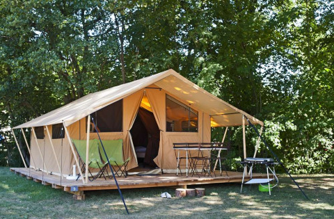 Una tienda tipo safari sobre una plataforma de madera en el bosque, con sillas, mesa y barbacoa delante.