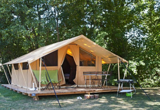 Una tienda tipo safari sobre una plataforma de madera en el bosque, con sillas, mesa y barbacoa delante.