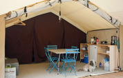 Intérieur de tente safari avec coin cuisine moderne, table et chaises bleues, et un petit chien sur le sol.