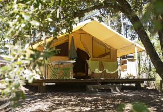A cozy safari tent with a porch, set among leafy trees, offering a comfortable glamping experience.
