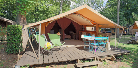 Tenda safari con terrazza in legno, sedie a sdraio verdi, tavolo blu, immersa in una zona alberata.