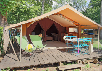 Tenda safari con terrazza in legno, sedie a sdraio verdi, tavolo blu, immersa in una zona alberata.