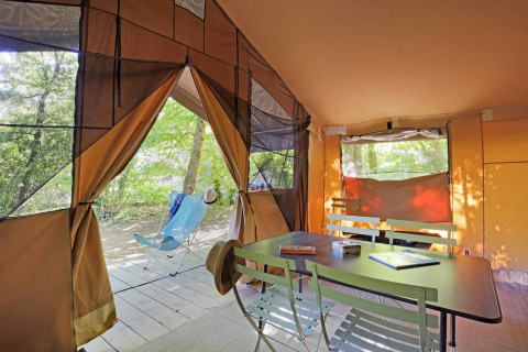 Interno di una tenda safari Toile&Bois tent Sweet con tavolo, sedie e vista sugli alberi fuori.
