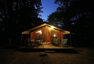 Tente safari Toile&Bois Sweet éclairée la nuit, terrasse en bois et sièges au cœur d’une forêt sombre.