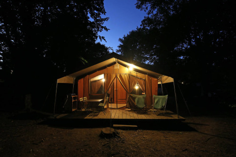 Safari-Zelt Toile&Bois Sweet am Abend, hell erleuchtet mit Holzterrasse und Stühlen im dunklen Wald.