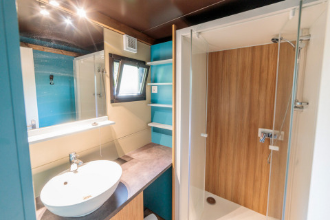 Baño moderno en Chalet Evasion glamping con lavabo redondo, ducha de madera y estantes azules.
