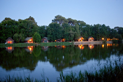 Acogedoras cabañas iluminadas se reflejan en el lago en Village Huttopia Lac de Rillé, Centre-Val de Loire, Francia.
