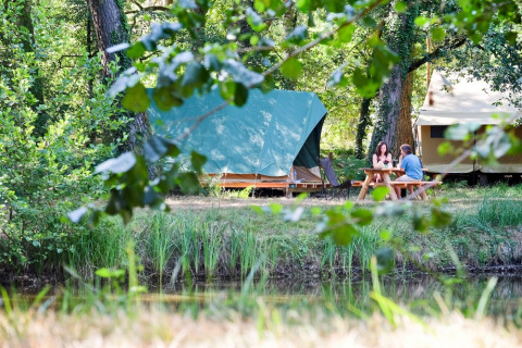 Dos personas sentadas en una mesa de picnic frente a tiendas en Village Huttopia Lac de Rillé, un parque en Francia.