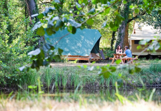 Due persone sedute a un tavolo da picnic davanti a tende nel verde di Village Huttopia Lac de Rillé, Francia.